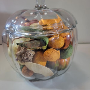 May include: A clear glass jar shaped like a pumpkin with a matching lid. The jar is filled with a variety of dried botanical elements, including orange, green, and brown textured pieces, seed pods, and dried leaves in assorted shapes and sizes.