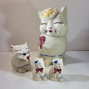 Puede incluir: Un juego de cuatro tarros de galletas de cerámica vintage con forma de gato. El tarro más grande presenta un gato blanco con un sombrero amarillo y un lazo rojo. Los otros tres tarros son más pequeños y presentan gatos blancos con lazos azules.