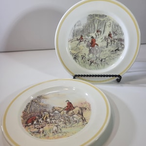 Puede incluir: Dos platos blancos con borde amarillo, que representan una escena de caza con jinetes a caballo, perros y árboles. Los platos se exhiben sobre una superficie blanca.