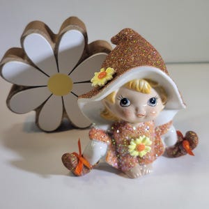 May include: A ceramic figurine of a seated fairy with a glittery orange dress and hat, adorned with yellow flower accents. The fairy has blonde hair and blue eyes. A large wooden daisy is in the background.