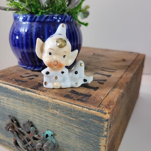 May include: A small white porcelain figurine of an elf with a gold hat and black polka dots, sitting on a wooden box with a blue painted drawer and a metal handle. The elf is wearing a white and gold outfit and has a black and white face.