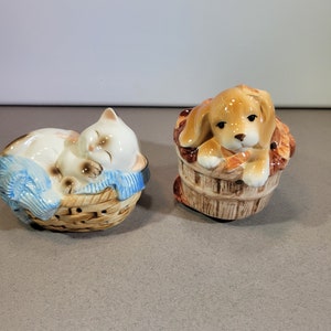 Puede incluir: Un juego de dos saleros y pimenteros de cerámica. El salero presenta un gato blanco durmiendo en una cesta de mimbre marrón con una manta azul. El pimentero presenta un perro marrón sentado en un cubo de madera marrón.
