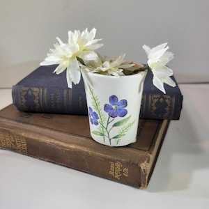 Puede incluir: Jarrón de cerámica blanco con un diseño floral con flores azules y hojas verdes. El jarrón está lleno de flores blancas.