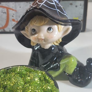 May include: A ceramic Halloween figurine of a witch elf with a black hat and outfit, and green shoes. The elf is kneeling near a cauldron filled with green glitter. The hat has a white spiderweb design.