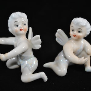 Puede incluir: Dos figuras de querubines de porcelana blanca con alas iridiscentes y rasgos faciales pintados. Los querubines están arrodillados y tienen los brazos extendidos.