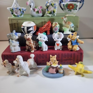 May include: A collection of miniature ceramic figurines including animals, a red glittery high heel, a small lidded pot, and a yellow airplane. The items are arranged on three stacked books, featuring various colors like white, blue, and brown.