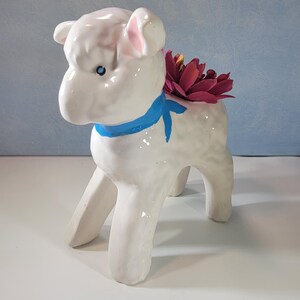 May include: A white ceramic lamb planter with a blue ribbon around its neck and a dark pink flower arrangement in its back. The lamb has blue eyes and pink inner ears. The planter is a decorative item for home decor.
