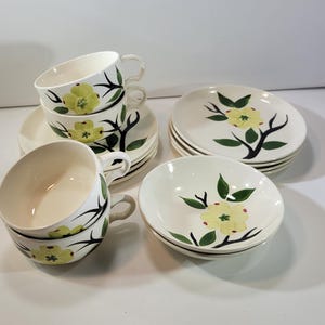 May include: A vintage cream-colored dish set featuring cups, saucers, and bowls. Each piece is adorned with a yellow floral design with green leaves and black branches. The set includes four cups, saucers, and bowls.