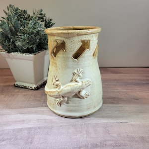 May include: A white ceramic vase with a brown glaze and a cutout design of a lizard. The vase has a speckled brown glaze and a cutout design of arrows around the top.