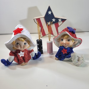 May include: Two ceramic elf figurines with red, white, and blue glitter clothing and hats. A wooden star with a patriotic design is behind them, along with decorative sticks in black and red-striped holders. The figurines have blonde hair and are in a sitting position.