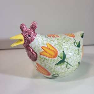 May include: A ceramic chicken ornament with a yellow beak and a red comb. The body is white with a green speckled pattern and orange tulip designs. A decorative item for the home.