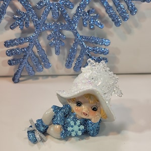 May include: A whimsical figurine of a child wearing a white hat and a blue glittery outfit, adorned with a snowflake. A large, glittery blue snowflake is in the background. The figurine is lying down, with a cheerful expression.