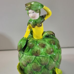 May include: A ceramic lidded jar shaped like an artichoke with a figure emerging from the top. The figure has a yellow torso, green leaf-like collar and hat, and a pale face. The base is a round, textured green artichoke with brown accents.