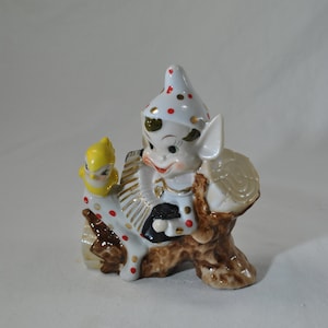 May include: A white ceramic figurine of an elf wearing a red and white polka dot hat, holding a small yellow bird and a brown accordion. The elf is sitting on a brown tree stump.