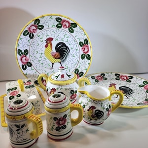 Puede incluir: Un conjunto de vajilla de cerámica con diseño de gallo y flores. Incluye platos, una tetera, una jarra de crema, una mantequera y saleros y pimenteros. Las piezas tienen un fondo blanco con detalles amarillos, rojos y verdes.