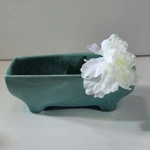 May include: A teal ceramic planter with a speckled finish, resting on four small feet. A large white flower with multiple petals is placed inside the planter, creating a decorative floral arrangement. The planter is empty.
