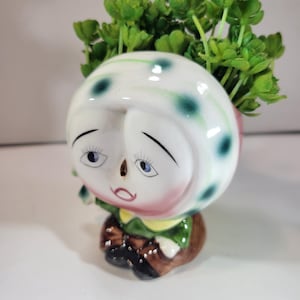 Puede incluir: Una maceta de cerámica caprichosa con forma de cabeza de personaje de dibujos animados, con una cara blanca, ojos azules, mejillas rosadas y un gorro de lunares verdes y blancos. La maceta está llena de plantas artificiales verdes.