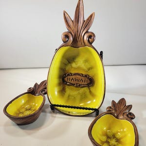 May include: A vintage ceramic pineapple-shaped serving set. The set includes a large serving dish and two smaller bowls, all with a yellow interior and brown exterior. The large dish has the word "HAWAII" in a brown, decorative font.