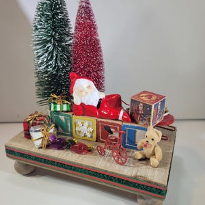 Puede incluir: Una exhibición navideña con una figura de Papá Noel, bloques de juguete, árboles de Navidad en miniatura y regalos envueltos. La exhibición incluye un pequeño triciclo rojo y un oso de peluche, todo dispuesto sobre una plataforma de madera con una cinta verde y roja.