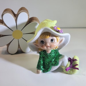 Puede incluir: Una figura de cerámica de un elfo sentado con una parte superior verde brillante, pantalones blancos y zapatos amarillos atados con cintas moradas. El elfo lleva un sombrero amarillo brillante con una flor morada. Una margarita de madera en el fondo.