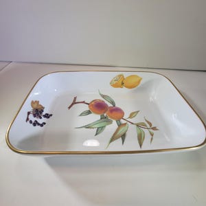 May include: White rectangular serving dish with a gold rim. The dish is decorated with a painted design of peaches, lemons, and grapes on a branch. The fruit is in shades of yellow, orange, purple, and green.
