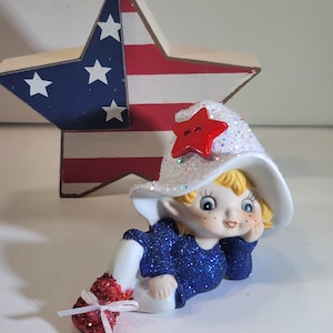 Puede incluir: Una figura patriótica de un elfo sonriente que lleva un atuendo azul brillante y un sombrero blanco con purpurina y una estrella roja. Una estrella de madera con un diseño de bandera americana pintada está en el fondo.