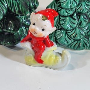 May include: A ceramic Christmas elf figurine wearing a red hat and outfit, seated on a yellow and white shoe. Two green ceramic Christmas trees are in the background.