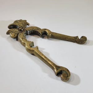 May include: A vintage brass nutcracker with an ornate floral design. The nutcracker features two handles and a central cracking mechanism. The brass has a warm, aged patina, suggesting its use over time. The overall design is decorative and functional.
