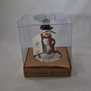 May include: A white snowman cookie press with a black top hat, red and green scarf, and a gold chain. The cookie press is in a clear plastic box with a brown cardboard base that says "cookie press".