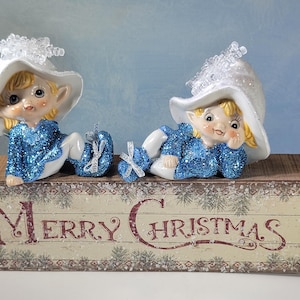 May include: Two elf figurines with blonde hair and blue glittery outfits wearing white hats with clear snowflake accents, positioned on a rectangular wooden block with a distressed finish that features the text Merry Christmas.