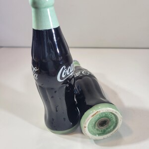 K&ouml;nnte beinhalten: Zwei Salz- und Pfefferstreuer aus Keramik in Form von Coca-Cola-Flaschen. Die Flaschen sind dunkelblau mit wei&szlig;em "Coca-Cola"-Schriftzug. Die Deckel sind hellgr&uuml;n mit blauen Kronkorken. Die Streuer stehen auf einer wei&szlig;en Oberfl&auml;che.