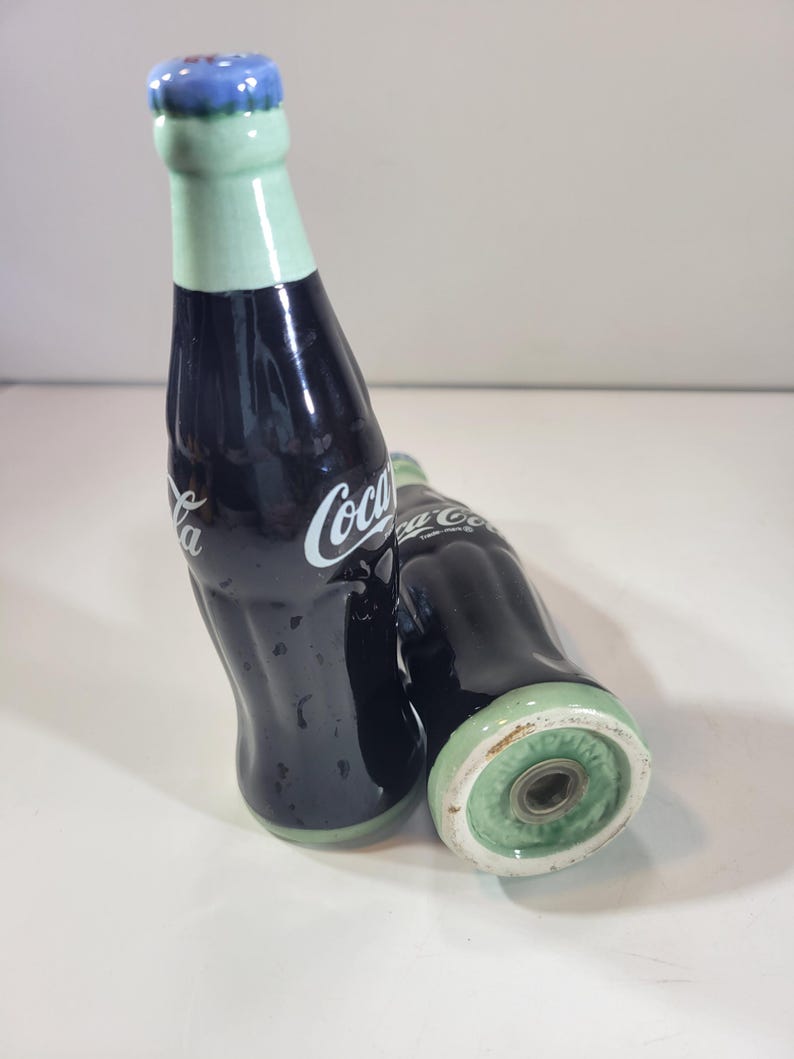 K&ouml;nnte beinhalten: Zwei Salz- und Pfefferstreuer aus Keramik in Form von Coca-Cola-Flaschen. Die Flaschen sind dunkelblau mit wei&szlig;em "Coca-Cola"-Schriftzug. Die Deckel sind hellgr&uuml;n mit blauen Kronkorken. Die Streuer stehen auf einer wei&szlig;en Oberfl&auml;che.