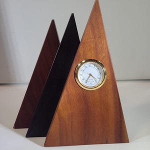 May include: Three wooden pyramid-shaped clocks in varying shades of brown and black. The front clock features a gold-rimmed clock face with white background and black numerals. The other two pyramids are solid colors.