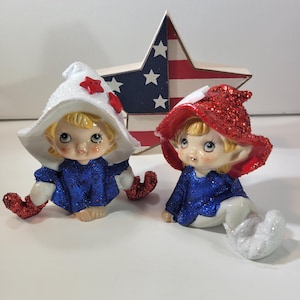 May include: Two ceramic figurines of elves in patriotic colors. One elf wears a white hat with a red star, the other a red hat. Both have blue glittery tops and red glittery feet. A wooden star with red, white, and blue stripes is in the background.