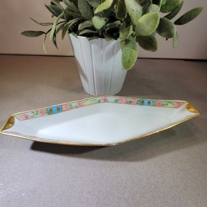 Puede incluir: Un plato de servir de porcelana blanca con un borde dorado y un diseño floral en rosa, azul y verde. El plato tiene forma de óvalo largo con extremos puntiagudos.