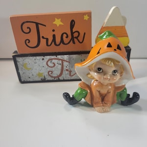 Puede incluir: Una figura de cerámica de un niño con un sombrero de calabaza y pantalones verdes, colocada frente a dos bloques de madera rectangulares apilados. El bloque superior es naranja con la palabra Trick en negro y el inferior tiene un diseño gris.