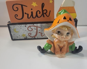 May include: A ceramic figurine of a child wearing a pumpkin hat and green pants, positioned in front of two stacked rectangular wooden blocks. The top block is orange with the word Trick in black, and the bottom block has a gray patterned design.