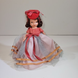 May include: A vintage porcelain doll wearing a red hat and a pink and gray tulle dress with gold trim.