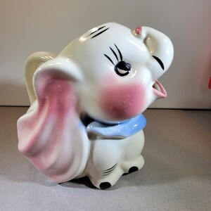 以下が含まれることがあります： ピンクの耳と青いリボンが付いた、象の形をした白いセラミックピッチャー。象は黒い目とピンクの鼻をしています。