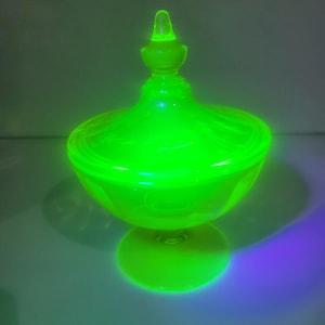 May include: A glowing green glass candy dish with a lid. The dish has a round bowl shape with a pedestal base and a decorative finial on the lid. The glass appears to be vintage and has a bright, neon-like appearance.