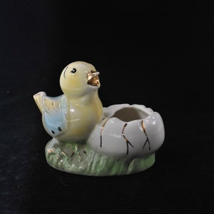 Puede incluir: Figura de cerámica de un pollito amarillo saliendo de un huevo blanco agrietado. El pollito tiene detalles azules y dorados. El huevo descansa sobre un trozo de hierba verde. Objeto decorativo.