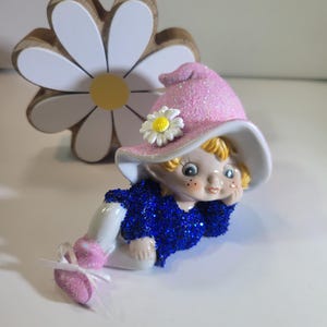 May include: A whimsical figurine of a fairy wearing a pink glittery hat with a white brim and a blue glittery top. The fairy is lying down, with white and pink shoes. A wooden daisy is in the background.