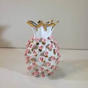 May include: A white ceramic pineapple-shaped vase with a gold-trimmed crown and pink rose accents. The pineapple body is covered in small, three-dimensional pink roses with gold detailing. The vase is a decorative item.