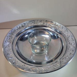 May include: A silver-colored serving tray with an embossed floral design around the rim. The tray has a recessed center that holds a clear glass tumbler. The tray is likely used for serving snacks or appetizers.