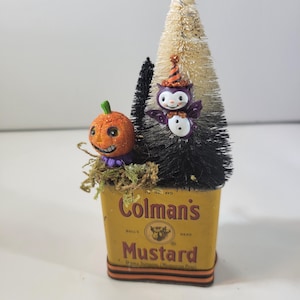 Puede incluir: Decoración de Halloween en una lata de mostaza Colman's. La lata amarilla muestra el texto "Colman's Mustard". En el interior hay una pequeña figura de calabaza naranja, una figura de búho en blanco y negro y dos árboles de cepillo. La calabaza y el búho tienen detalles festivos.