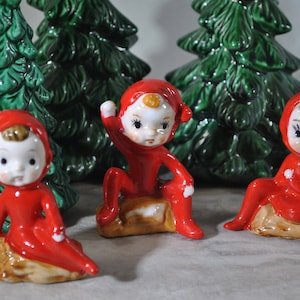 May include: Three ceramic elf figurines in red outfits with white faces and brown hair. The elves are posed on small, brown bases, with two green ceramic Christmas trees behind them. These figurines are likely vintage Christmas decorations.