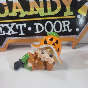 May include: A ceramic figurine of a reclining elf wearing an orange pumpkin hat with black triangular eyes and a brown outfit with green pants. Behind the figurine is a black sign with white polka dots and the text CANDY NEXT DOOR.