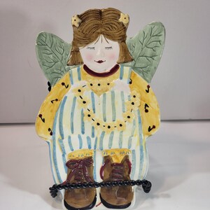 May include: A ceramic angel figurine with brown hair, closed eyes, and a smiling face. The angel wears a yellow and blue striped outfit with brown boots. Green leaf-shaped wings are visible on the back. A black rope is wrapped around the boots.