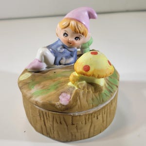 Puede incluir: Una caja de baratijas de cerámica con una figura de gnomo en una chaqueta azul y un sombrero rosa, descansando sobre una tapa en forma de hongo. La caja es marrón con un diseño de corteza texturizada, y el hongo es amarillo con puntos rojos.
