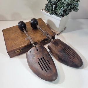 Puede incluir: Un par de hormas de zapatos de madera con reguladores metálicos y pomos negros. Las hormas están en una caja de madera.
